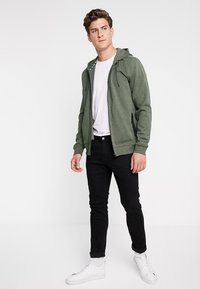 Groene hoodie met rits en verstelbare trekkoorden, gedragen over een wit t-shirt en zwarte broek, gecombineerd met witte sneakers. Zachte stofstructuur.