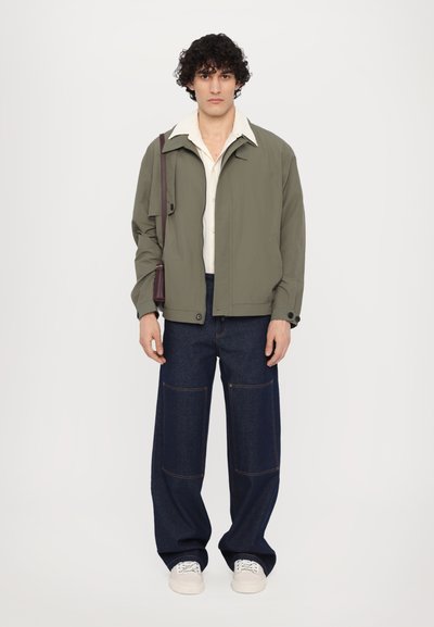 Olivengrønn jakke med krage, over en kremfarget skjorte med knapper. Løse mørk denimbukser med kontrastsøm og hvite joggesko.