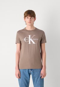 Ruda medvilninė marškinė su trumpomis rankovėmis, ant kurių pavaizduotas didelis baltas "CK" logotipas ir tekstas "Calvin Klein Jeans". Dėvima su mėlynais džinsais.