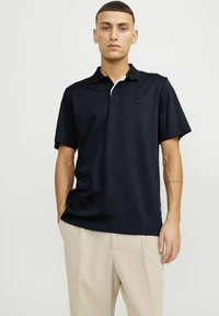 Mornarska polo majica s kratkimi rokavi temno modre barve z kontrastnim belim ovratnikom, lahka tkanina, sproščen kroj in subtilen logotip na prsih.