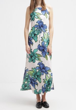 Vrouw draagt een witte mouwloze maxi-jurk met grote blauw-groene bloemenprint en zwarte instapschoenen.
