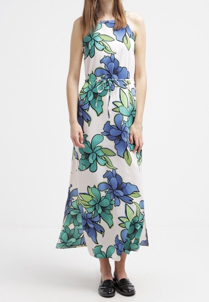 Femme portant une robe maxi blanche sans manches avec un grand imprimé floral bleu et vert et des chaussures noires sans lacets.