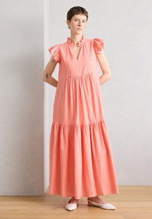 Kvinde med kort hår står mod en ensfarvet væg iført en lang koralrosa kjole med flæset korte ærmer og hvide spidse flade sko.