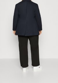 Blazer bleu marine à la coupe ajustée, avec une fente arrière, porté sur un pantalon noir et des baskets blanches avec un logo. Texture de tissu lisse.