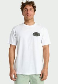 Hombre vestido con una camiseta de manga corta blanca con un pequeño gráfico circular negro en el lado izquierdo del pecho y pantalones verdes claros, de pie frente a un fondo sencillo.