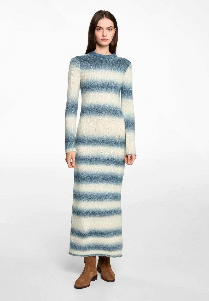 Pitkä neulemekko, jossa on sinisiä ja vaaleankirjavia gradienttiraitoja, pyöreä pääntie ja pitkät hihat, yhdistettynä ruskeisiin nilkkureihin.