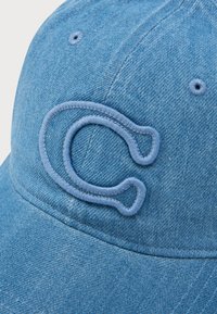 Denim kapa v svetlo modri barvi z teksturirano površino. Na sprednji strani ima veliko, vezeno črko "C" v usklajeni barvi niti.