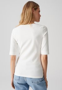 Vrouw met blond haar die een eenvoudig wit top met ellebooglange mouwen en lichtblauwe spijkerbroek draagt, gezien van achteren tegen een grijze achtergrond.