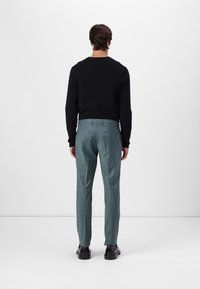 Suéter negro combinado con pantalones en tono verde azulado que presentan un corte ajustado, con detalles en los bolsillos traseros, y zapatos negros, visto desde atrás.