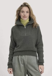 Dunkelgrüner Pullover mit hohem Kragen und einem halben Reißverschluss, der eine gerippte Textur am Hals aufweist. Kombiniert mit hellgrünen gestreiften Hosen.