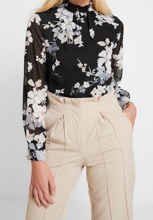 Svart blommig blus med långa genomskinliga ärmar som har ljusa blommor, en hög krage med smock, i kombination med beiga plisserade byxor.