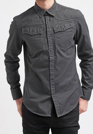 Homme portant une chemise gris foncé cintrée avec des poches à rabat sur la poitrine et des manches longues, debout les mains dans les poches.