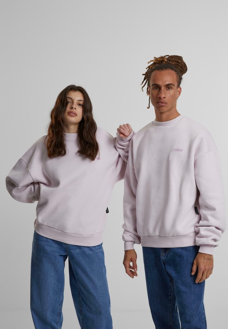 Lys rosa hettegensere med rund hals, oversized passform og ribbede mansjetter, kombinert med blå jeans. En hettegenser har et lite logo.