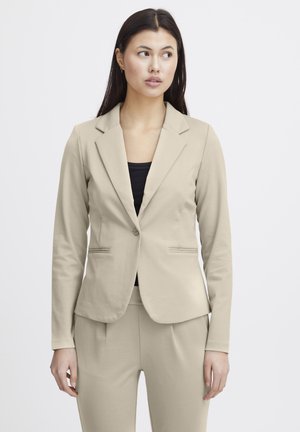 Femeie purtând un blazer bej croită și pantaloni asortați peste un top negru, uitându-se ușor spre stânga pe un fundal simplu.