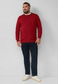 Rode gebreide trui met een ronde hals, gecombineerd met donkerblauwe jeans en witte sneakers. De stof is glad met geribde manchetten en zoom.