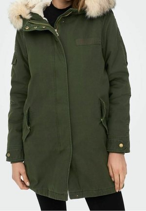 Femme portant un long manteau parka vert olive à capuche bordée de fourrure et poignets de manches boutonnés, mains le long du corps.