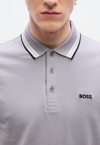 Γκρι πόλο μπλούζα  με υφή ύφανσης, δύο κουμπιά και μαύρο κεντημένο λογότυπο "BOSS". Διαθέτει γιακά με λευκές και μαύρες ρίγες.