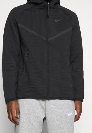 Schwarzer Nike Kapuzenpullover mit Reißverschluss, strukturiertem Stoff und Seitentaschen, getragen mit hellgrauen Nike Jogginghosen.