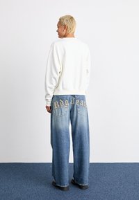 Sudadera blanca con cuello redondo combinada con unos vaqueros de corte suelto y desgastados de color azul, que presentan bordado "Bdg Jeans" en los bolsillos traseros.