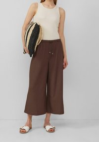 Svetle béžová tílko, hnědé volné kalhoty s tkaničkou v pase a bílé klouzavé sandály s mašlí. Drží pruhovanou pletenou tašku.