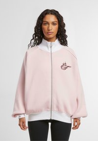 Felpa rosa con zip e dettagli bianchi, vestibilità rilassata e polsini a coste. Presenta il logo sul lato sinistro del petto e una texture liscia e morbida.