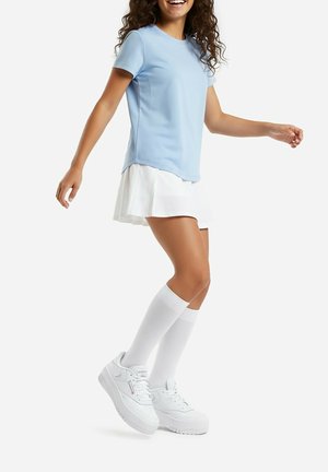 Mujer con camiseta azul claro, falda plisada blanca, calcetas blancas hasta la rodilla y zapatillas blancas de plataforma, de pie con los brazos ligeramente levantados.