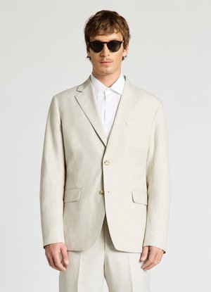 Homme portant un costume beige avec une chemise blanche et des lunettes de soleil foncées, debout devant un fond clair uni.