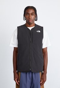 Fekete párnázott mellény, elől cipzárral, magas nyakkivágással és a bal mellen a The North Face logójával. Fehér póló fölött viselve.