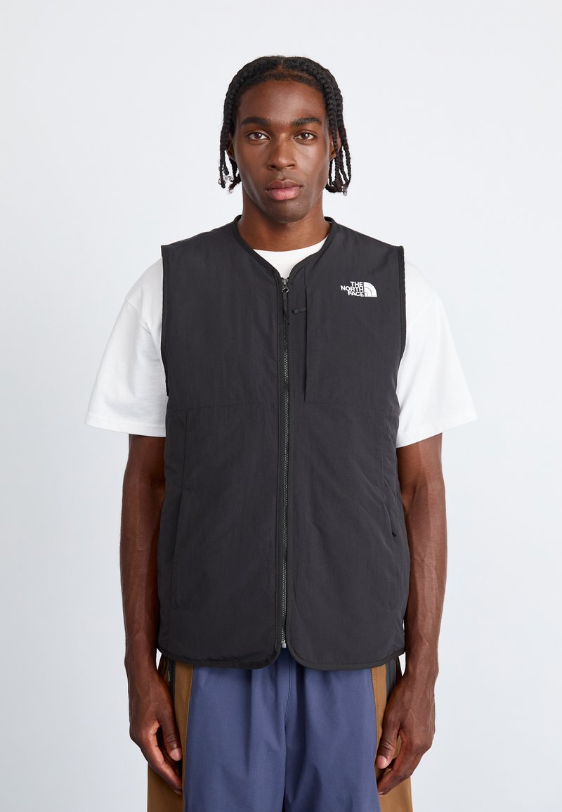 Fekete párnázott mellény, elől cipzárral, magas nyakkivágással és a bal mellen a The North Face logójával. Fehér póló fölött viselve.