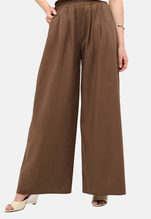 Personne portant un pantalon marron taille haute à jambes larges et des sandales à talons crème, debout une main sur la hanche devant un fond uni.