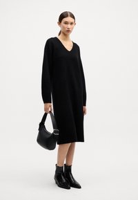 Rochie tricotată neagră cu decolteu în V și mâneci lungi, asortată cu o geantă mică neagră și cizme negre cu vârf ascuțit până la gleznă.