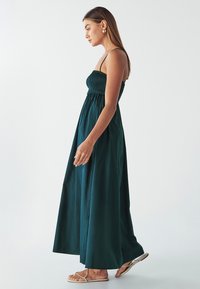 Abito lungo senza maniche di colore verde, realizzato in cotone. Presenta un corpetto arricciato e una gonna fluida. Il modello indossa semplici sandali appiatti.