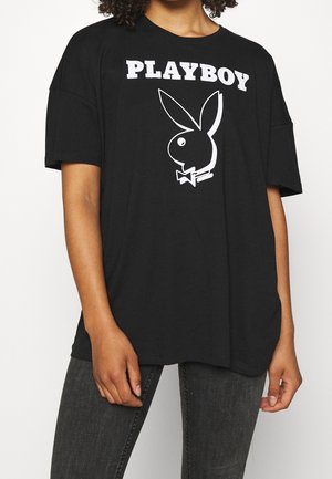 Persoon draagt een losse zwarte t-shirt met witte "PLAYBOY"-tekst en een groot konijnenlogo, gecombineerd met donkergrijze spijkerbroek.