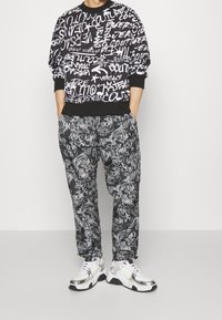 Černá mikina s bílým textem ve stylu graffiti, spárovaná s černými kalhotami s vířícími vzory a teniskami se stříbrnou podrážkou.