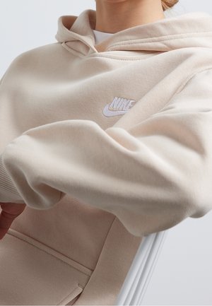 Sudadera con capucha beige de textura suave, con un bolsillo frontal y un logo blanco de Nike en el pecho. Incluye una capucha con cordón ajustable.