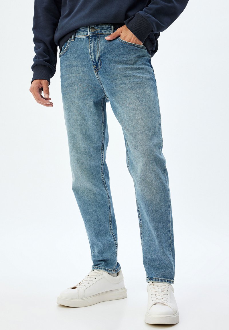 Jeans in denim azzurro chiaro con taglio dritto, cinque tasche, chiusura con bottone e zip, e una leggera scoloritura per texture. Abbinati a sneakers bianche.