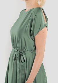 Grön satinklänning med lös passform, korta ärmar och knyt i midjan. Har öppna axellinningar och en flödande kjoldesign.