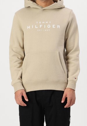 Mężczyzna ubrany w beżową bluzę z kapturem Tommy Hilfiger z przednią kieszenią i nadrukowanym logo, w połączeniu z czarnymi spodniami, stojący na białym tle.