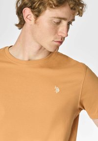 T-shirt a maniche corte con scollo rotondo in cotone arancione bruciato, con un sottile logo ricamato in oro. Texture liscia e vestibilità rilassata.