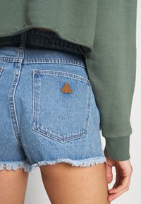 Denim shorts in light blue with frayed hems, featuring a brown triangular leather patch on the back pocket and a green long-sleeve top.