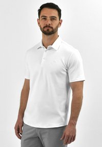 Witte polo met een drieknops sluiting, zachte textuur, korte mouwen en een subtiele logo op de borst, gecombineerd met lichtgrijze shorts.