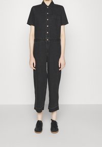 Svart denim jumpsuit med korta ärmar, knappstängning fram, bröstficka och cropped ben. Har kontrasterande sömmar och en avslappnad passform.