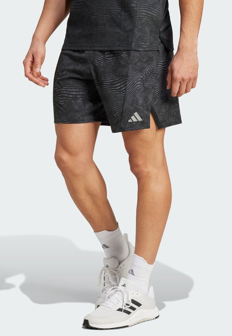 Crne atletske kratke hlače s uzorkom valova, bočnim prorezima i Adidas logotipom na donjem lijevom dijelu. Uparene s bijelim tenisicama i čarapama.