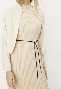 Giacca bomber crema sopra un vestito beige plissettato. Il vestito ha un collo alto e una cintura nera regolabile in vita. Tessuti morbidi in tutto.