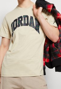 Beżowa koszulka z krótkim rękawem z czarno-zielonym logo "JORDAN" w kratkę. Noszona z czarną i czerwoną kurtką w kratkę. Model trzyma kurtkę.