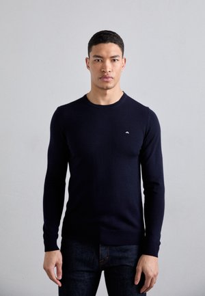 Maglione lungo a maniche, di colore blu navy, con un design aderente, scollatura rotonda e polsini a costine. Piccolo dettaglio del logo sul petto, realizzato in materiale morbido.