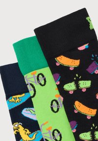 Τρεις πολύχρωμες κάλτσες με σχέδια κίτρινων δεινοσαύρων σε μπλε σανίδες σερφ, μοβ ποδήλατα σε πράσινο φόντο και πορτοκαλί και πράσινες σανίδες σκέιτμπορντ σε μαύρο φόντο.