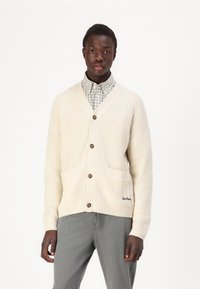 Cárdigan de punto color crema con cuello en V, tres botones, dos bolsillos frontales y logo bordado. Llevado sobre una camisa a cuadros en gris y blanco.