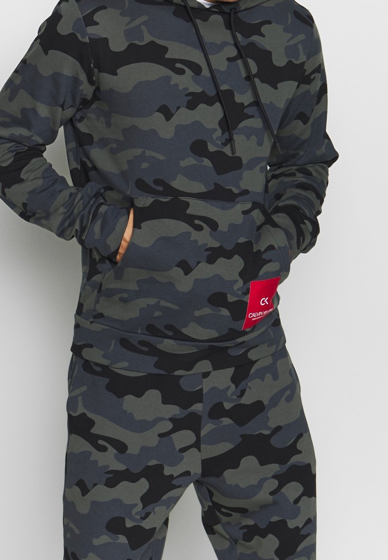 Sweatshirt à capuche et pantalon assorti en motif camouflage gris foncé. Comprend une poche avant et un patch logo rouge sur la ceinture.