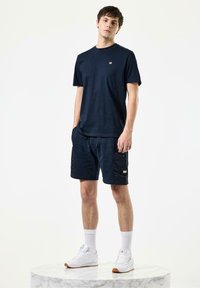 Marineblå bomulds t-shirt med korte ærmer og lille logo, som er parret med marineblå shorts med en cargo-lomme og hvide sneakers. Glat tekstur.